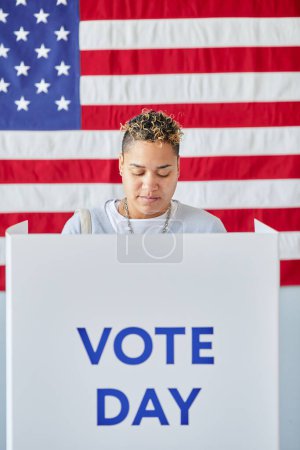 Téléchargez les photos : Portrait vertical de la jeune femme noire debout dans l'isoloir le jour du scrutin contre le drapeau américain - en image libre de droit