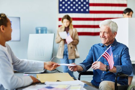 Téléchargez les photos : Portrait d'une personne âgée souriante handicapée recevant un bulletin de vote au bureau de vote le jour du scrutin avec drapeau américain en arrière-plan - en image libre de droit