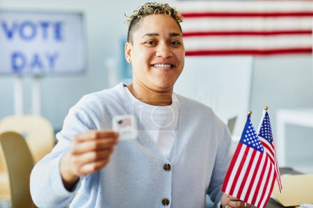 Foto de Retrato de la cintura hacia arriba de una persona sonriente sosteniendo la etiqueta de votación y la bandera estadounidense el día de las elecciones - Imagen libre de derechos