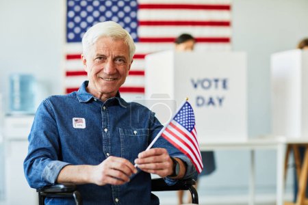 Téléchargez les photos : Portrait en première vue d'un homme âgé souriant handicapé tenant le drapeau des États-Unis dans un bureau de vote, espace de copie - en image libre de droit