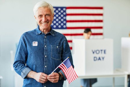 Téléchargez les photos : Portrait taille haute d'un homme âgé souriant tenant le drapeau américain dans un bureau de vote le jour du scrutin, espace de copie - en image libre de droit