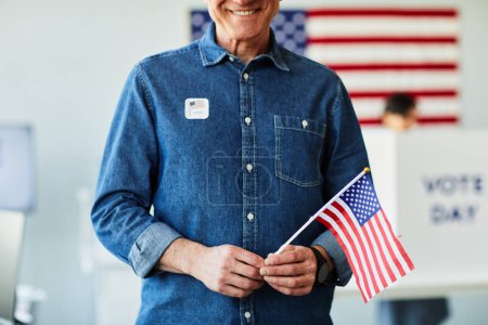 Téléchargez les photos : Portrait recadré d'un homme âgé souriant tenant le drapeau américain dans un bureau de vote le jour du scrutin, espace de copie - en image libre de droit