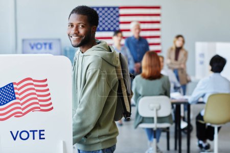 Téléchargez les photos : Portrait latéral d'un homme noir souriant votant en cabine le jour du scrutin et regardant la caméra, l'espace de copie - en image libre de droit
