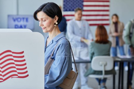 Téléchargez les photos : Portrait latéral d'une élégante femme adulte votant en cabine le jour du scrutin avec des drapeaux américains en plan, espace de copie - en image libre de droit