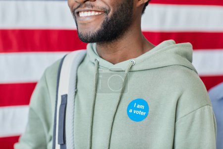 Téléchargez les photos : Gros plan de l'homme noir souriant avec je suis un autocollant de l'électeur contre drapeau américain en arrière-plan, espace de copie - en image libre de droit