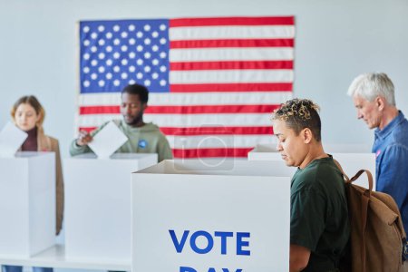 Téléchargez les photos : Groupe diversifié de personnes votant dans des isoloirs au bureau électoral avec drapeau américain, espace de copie - en image libre de droit