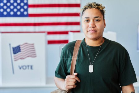 Téléchargez les photos : Portrait taille haute de la femme noire moderne regardant la caméra dans le bureau de vote contre le drapeau américain, espace de copie - en image libre de droit