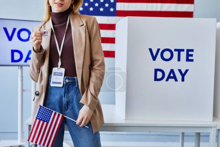 Téléchargez les photos : Plan recadré d'une jeune femme tenant un autocollant I vote debout dans un bureau de vote avec drapeau américain, espace de copie - en image libre de droit