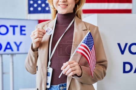 Téléchargez les photos : Portrait recadré d'une jeune femme souriante tenant un autocollant de vote et un drapeau américain debout dans un bureau de vote - en image libre de droit