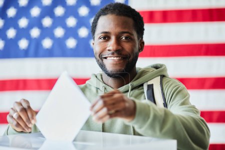 Téléchargez les photos : Portrait d'un homme noir souriant mettant son bulletin de vote à la poubelle contre le drapeau américain - en image libre de droit