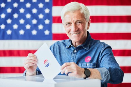 Foto de Retrato de hombre mayor sonriente poniendo papeleta en la papeleta contra la bandera estadounidense, espacio para copiar - Imagen libre de derechos