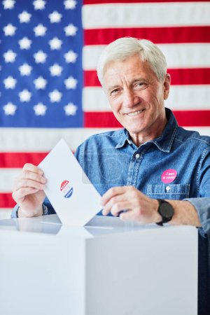 Téléchargez les photos : Portrait vertical d'un homme âgé souriant mettant son bulletin de vote en poubelle contre le drapeau américain en arrière-plan - en image libre de droit