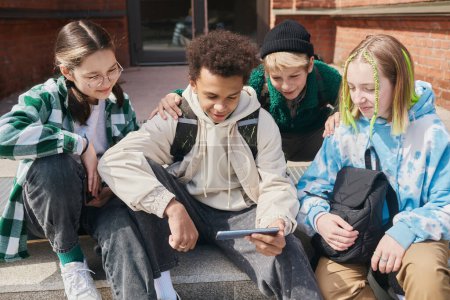 Téléchargez les photos : Groupe d'adolescents assis sur les marches de leur école en utilisant un smartphone pour regarder du contenu vidéo après les cours - en image libre de droit