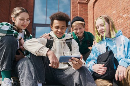 Téléchargez les photos : Groupe d'écoliers positifs regardant des vidéos sur smartphone et souriant assis à l'extérieur - en image libre de droit