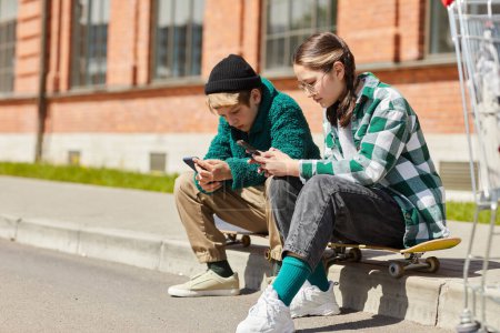 Téléchargez les photos : Couple d'adolescents assis n leurs planches à roulettes à l'extérieur et bavarder en ligne à l'aide de smartphones - en image libre de droit