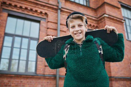 Téléchargez les photos : Portrait d'un adolescent tenant un skateboard souriant à la caméra tout en marchant à l'extérieur - en image libre de droit