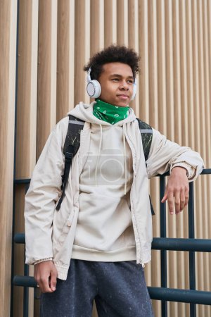 Photo for African teenage boy in casual clothing with backpack listening to music in wireless headphones standing outdoors - Royalty Free Image