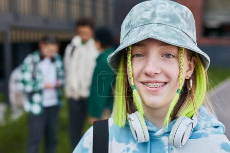 Foto de Retrato de adolescente en sombrero con el pelo teñido de color sonriendo a la cámara de pie al aire libre con sus amigos de fondo - Imagen libre de derechos