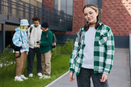 Portrait d'adolescente en lunettes regardant la caméra debout dans la cour de l'école avec ses amis en arrière-plan