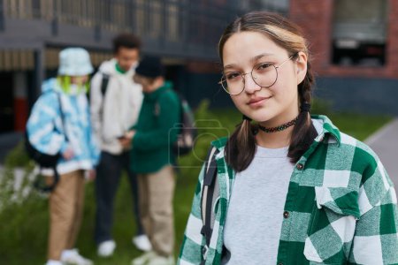 Foto de Retrato de una joven colegiala con anteojos y camisa de cuadros mirando a la cámara mientras estaba de pie afuera con sus compañeros de clase en el fondo - Imagen libre de derechos