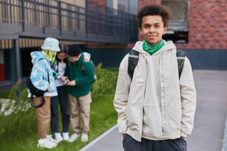 Téléchargez les photos : Portrait d'un adolescent africain avec un sac à dos debout avec les mains dans les poches et regardant la caméra avec des amis en arrière-plan - en image libre de droit