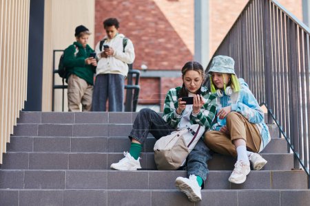 Photo for Two teenage girls sitting on stairs outdoors and watching video on mobile phone with young boys in background - Royalty Free Image