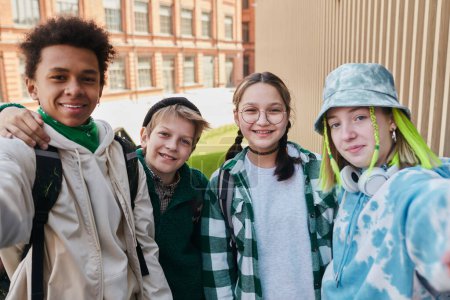 Téléchargez les photos : Portrait d'amis adolescents souriant à la caméra tout en faisant un portrait selfie sur smartphone debout à l'extérieur avec le bâtiment de l'école en arrière-plan - en image libre de droit