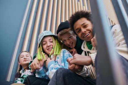 Foto de Retrato de amigos adolescentes cercanos felices sonriendo a la cámara mientras están sentados en los escalones al aire libre y abrazándose - Imagen libre de derechos