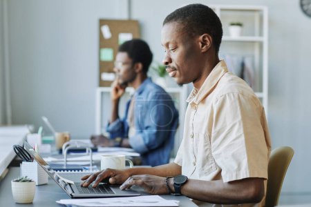 Téléchargez les photos : Portrait latéral d'un homme noir adulte utilisant un ordinateur pendant qu'il travaillait dans un bureau ou un espace de coworking - en image libre de droit