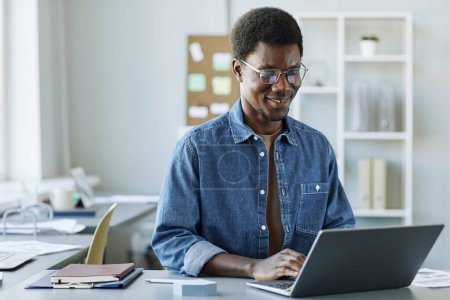 Téléchargez les photos : Portrait d'un homme noir souriant travaillant au bureau et utilisant un ordinateur portable dans un intérieur gris minimal, espace de copie - en image libre de droit