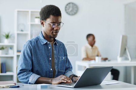 Téléchargez les photos : Portrait de jeune homme afro-américain travaillant au bureau et utilisant un ordinateur portable dans un intérieur gris minimal, espace de copie - en image libre de droit
