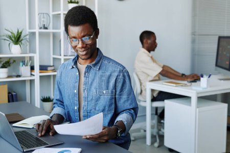 Téléchargez les photos : Portrait de jeune homme noir portant des lunettes analysant des documents tout en travaillant au bureau, espace de copie - en image libre de droit