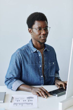 Téléchargez les photos : Portrait vertical de jeune professionnel noir sur le lieu de travail au bureau à l'aide d'un ordinateur PC contre un mur blanc - en image libre de droit