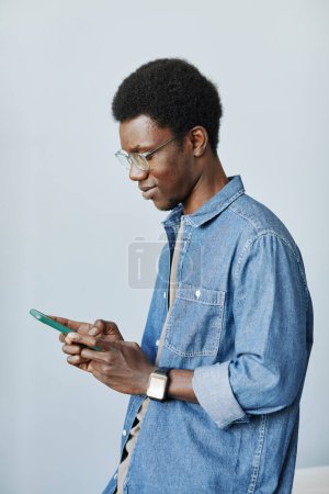 Photo for Vertical waist up portrait of young black man holding smartphone and typing messages against minimal grey background - Royalty Free Image