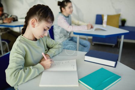 Foto de Retrato de vista lateral de una colegiala joven escribiendo en un cuaderno mientras está sentada en el escritorio durante la clase, espacio para copiar - Imagen libre de derechos