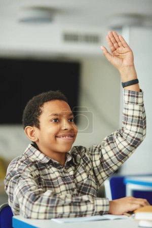 Téléchargez les photos : Portrait vertical de jeune garçon noir levant la main en classe et souriant joyeusement - en image libre de droit