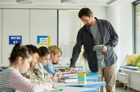 Photo for Side view portrait of male teacher helping children taking test in school classroom - Royalty Free Image