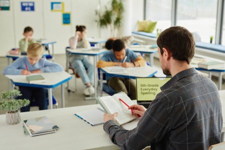 Téléchargez les photos : Vue de dos à l'enseignant mâle assis au bureau dans la salle de classe avec un groupe d'enfants passant le test en arrière-plan, espace de copie - en image libre de droit