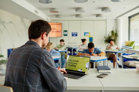 Téléchargez les photos : Vue de dos à l'enseignant masculin en utilisant un ordinateur portable au bureau dans la salle de classe avec un groupe d'enfants étudiant en arrière-plan, espace de copie - en image libre de droit