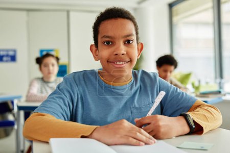 Foto de Retrato de vista frontal del colegial afroamericano mirando la cámara mientras está sentado en el escritorio en el aula de la escuela - Imagen libre de derechos
