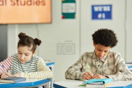 Photo for Front view at two children sitting at desk in classroom and writing in notebooks while studying in school - Royalty Free Image
