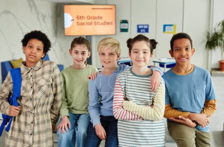 Téléchargez les photos : Portrait d'un groupe diversifié d'enfants regardant la caméra et souriant tout en restant ensemble dans la salle de classe de l'école - en image libre de droit