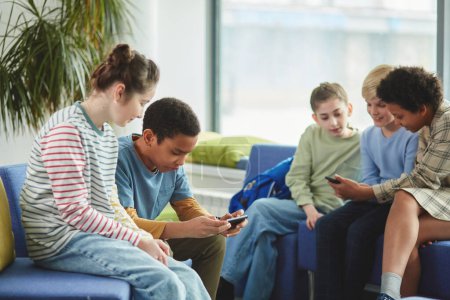 Téléchargez les photos : Groupe diversifié d'enfants utilisant des smartphones à l'école pendant la pause tous habillés en vêtements colorés - en image libre de droit