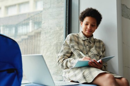 Téléchargez les photos : Portrait d'un enfant afro-américain souriant écrivant dans un cahier assis sur une fenêtre à l'école avec un ordinateur portable, espace de copie - en image libre de droit