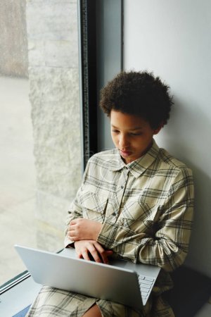 Téléchargez les photos : Portrait vertical de jeune fille noire à l'aide d'un ordinateur portable et regarder des vidéos tout en étant assis par la fenêtre à l'école - en image libre de droit