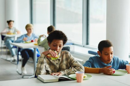 Photo for Portrait of schoolchildren at lunch break, focus on young african American girl eating sandwich and reading book, copy space - Royalty Free Image