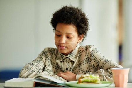 Téléchargez les photos : Vue de face portrait de jeune afro-américaine livre de lecture pendant la pause déjeuner dans la cantine scolaire - en image libre de droit