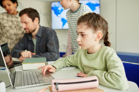 Téléchargez les photos : Vue de côté portrait d'écolière mignonne en utilisant un ordinateur portable dans la classe de l'école moderne avec des enfants en arrière-plan - en image libre de droit