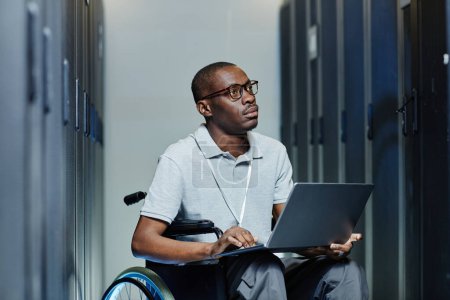 Foto de Retrato mínimo del joven negro con discapacidad que usa el portátil en la sala de servidores mientras trabaja como técnico de TI - Imagen libre de derechos