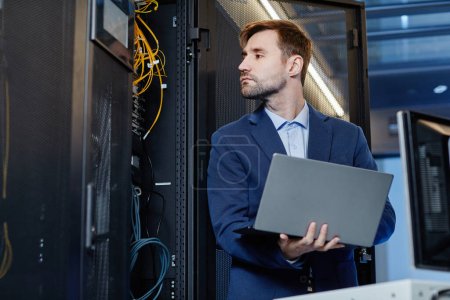 Photo for Waist up portrait of Caucasian young man holding laptop while setting up internet network in server room - Royalty Free Image